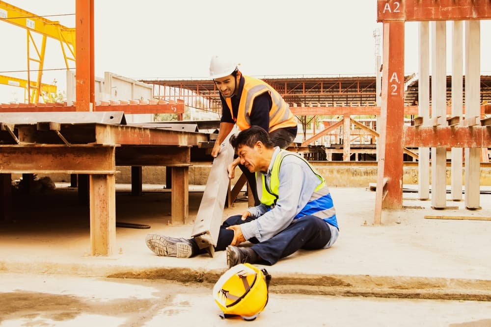 Injured construction worker holding his leg while a coworker helps at a job site