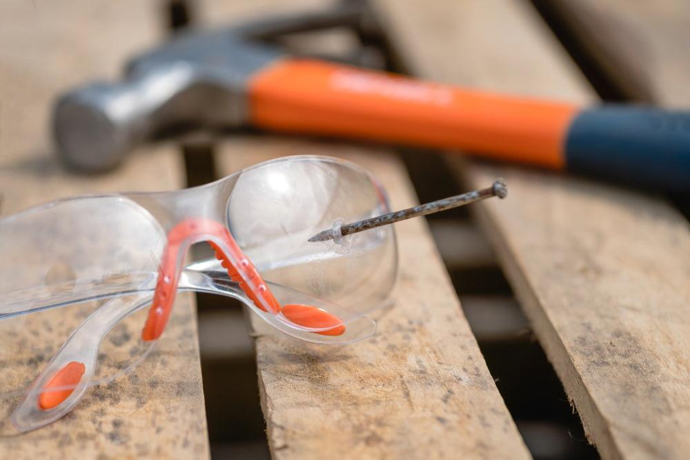 Nail piercing clear safety glasses beside a hammer on wooden boards